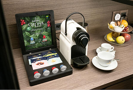 Cutie cu mostre de ceai și capsule de cafea Dallmayr lângă un aparat de cafea cu capsule în zona pentru mic dejun a hotelului